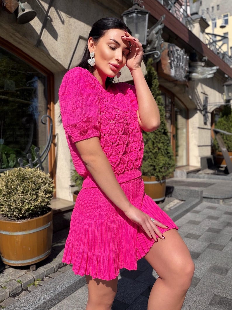 May include: A bright pink knitted two-piece outfit. The top has short sleeves and a detailed openwork pattern on the bodice. The matching skirt is a mini-skirt with a pleated hem. The outfit is made of a textured knit fabric.