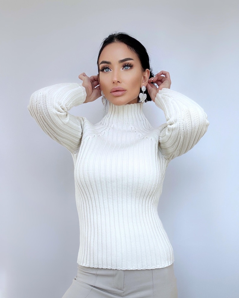 May include: A white ribbed turtleneck sweater with long sleeves. The sweater is fitted and has a high neckline.