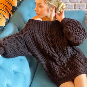 May include: A dark brown, off-the-shoulder, long-sleeved sweater featuring a chunky cable knit pattern. The garment has a wide, ribbed neckline and ribbed cuffs, with a thick, textured weave throughout the body and sleeves.