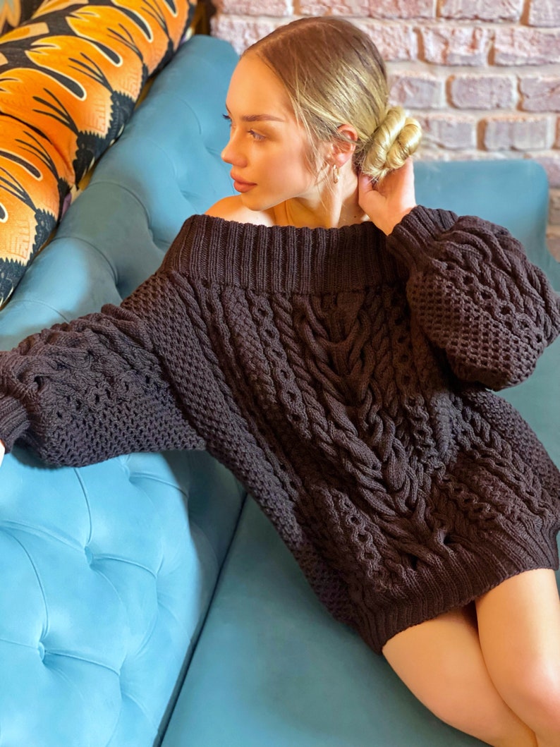 May include: A dark brown, off-the-shoulder, long-sleeved sweater featuring a chunky cable knit pattern. The garment has a wide, ribbed neckline and ribbed cuffs, with a thick, textured weave throughout the body and sleeves.