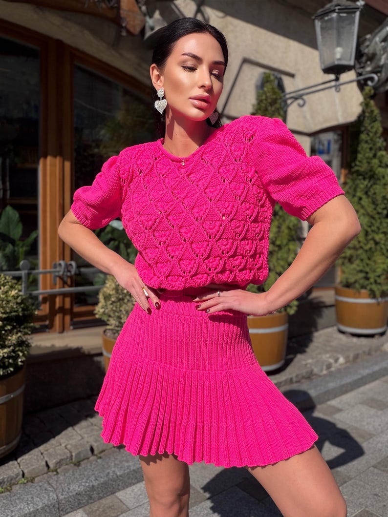 May include: A bright pink knit two-piece set. The top is a short-sleeved sweater with a textured diamond pattern. The matching skirt is pleated. The outfit is worn with heart-shaped earrings. The set is a vibrant and eye-catching ensemble.