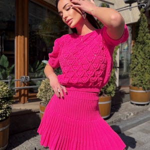 May include: A bright pink knit two-piece set. The top is a short-sleeved sweater with a textured pattern. The matching skirt is a mini skirt with a pleated design. The outfit is made of a soft, textured material.