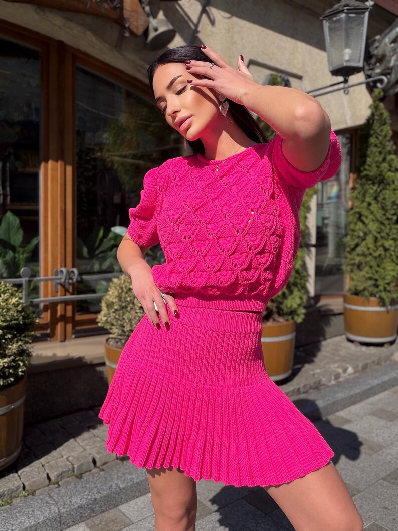 May include: A bright pink knit two-piece set. The top is a short-sleeved sweater with a textured pattern. The matching skirt is a mini skirt with a pleated design. The outfit is made of a soft, textured material.