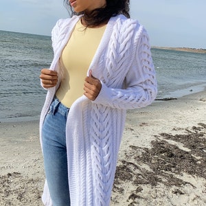 May include: A long, white cable-knit cardigan with a flowing design. The cardigan is worn over a yellow top and blue jeans. The person is wearing sunglasses and standing on a sandy beach near the water.