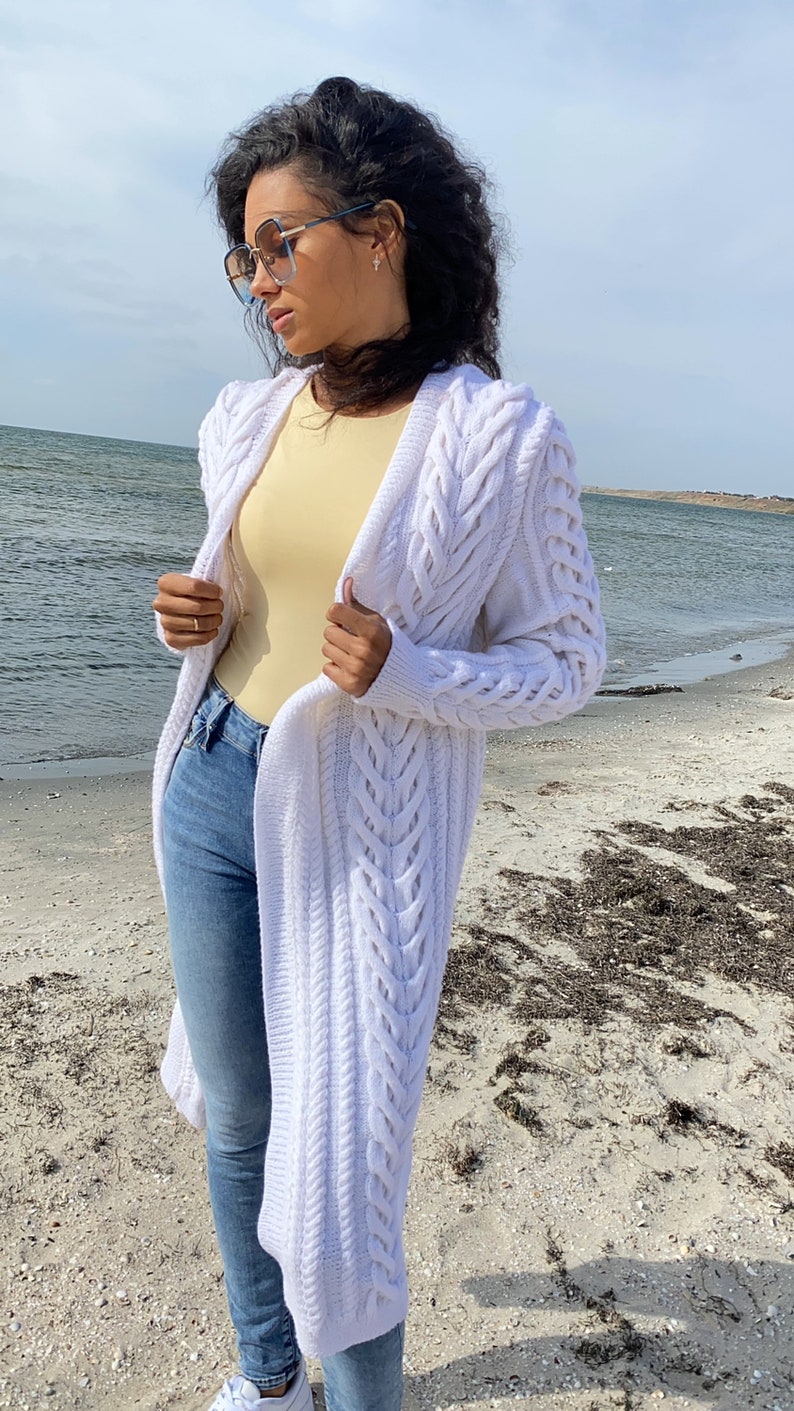 May include: A long, white cable-knit cardigan with a flowing design. The cardigan is worn over a yellow top and blue jeans. The person is wearing sunglasses and standing on a sandy beach near the water.