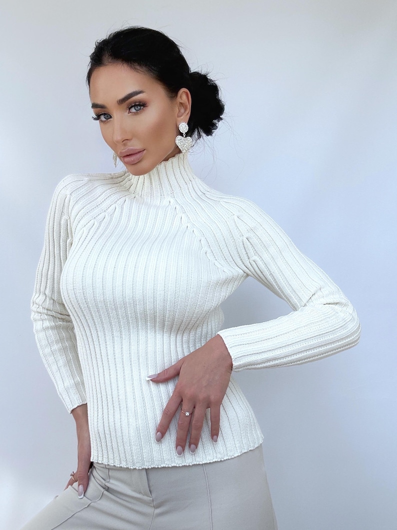 May include: A white ribbed turtleneck sweater with long sleeves. The sweater is fitted and has a slightly cropped length.