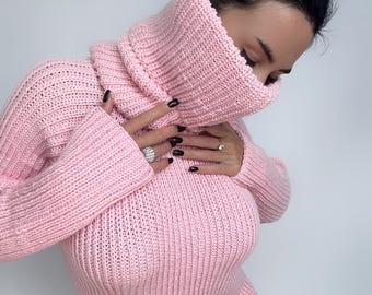 Puede incluir: Un suéter de manga larga en color rosa claro con una textura de punto grueso y un cuello alto de tortuga de gran tamaño que se extiende hacia arriba para cubrir la parte inferior del rostro. La prenda presenta un patrón acanalado en toda ella.