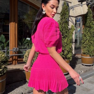 May include: A bright pink, two-piece outfit. The top has short, puffy sleeves and a textured fabric. The skirt is pleated and falls above the knees. The outfit is worn outdoors, with a background of greenery and a building.
