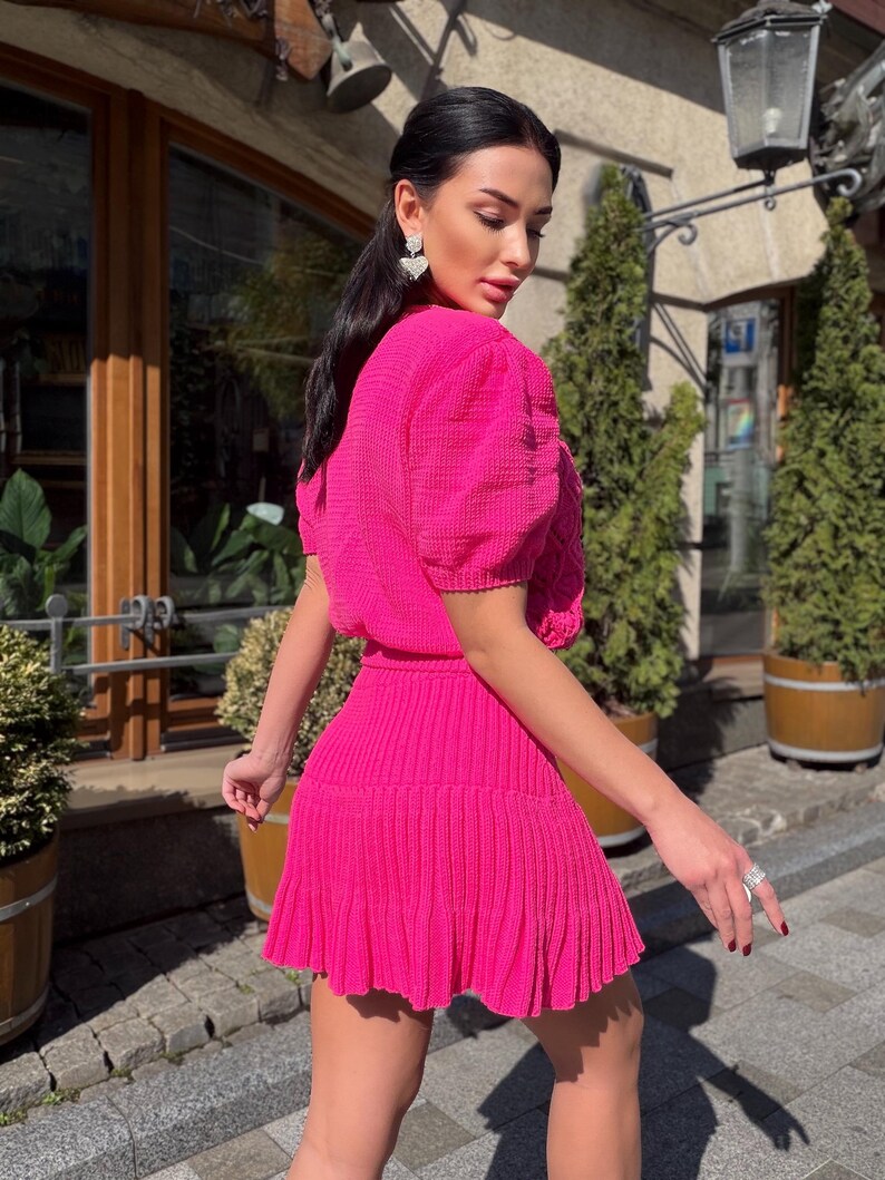 May include: A bright pink, two-piece outfit. The top has short, puffy sleeves and a textured fabric. The skirt is pleated and falls above the knees. The outfit is worn outdoors, with a background of greenery and a building.