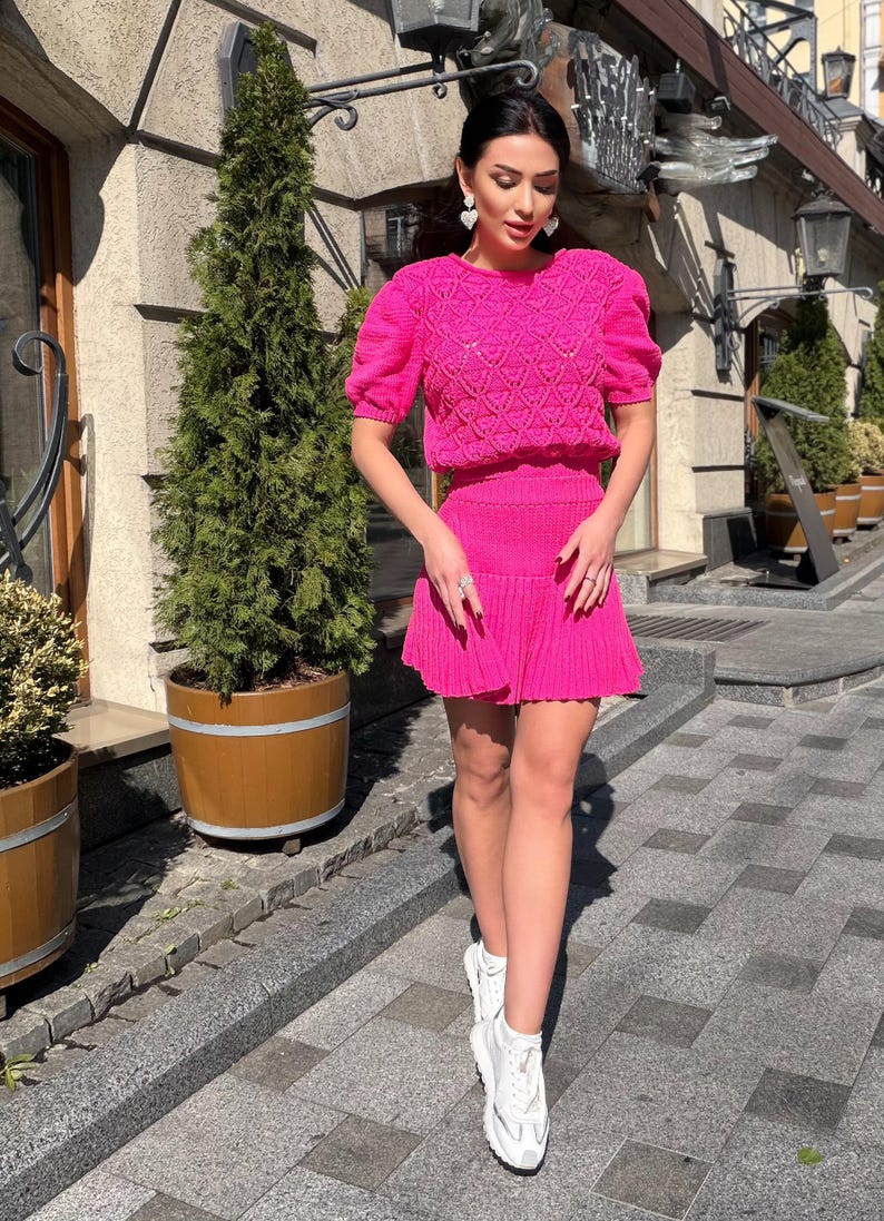 May include: A bright pink knit two-piece set. The top has short, puffed sleeves and a textured pattern. The matching skirt is pleated. The outfit is paired with white sneakers. The background includes a building and potted plants.