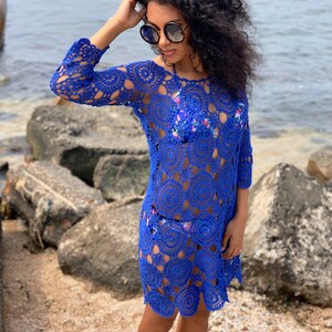 K&ouml;nnte beinhalten: Ein k&ouml;nigsblaues H&auml;kel-Strandkleid mit einem kreisf&ouml;rmigen Muster. Das Kleid hat lange &Auml;rmel und eine lockere Passform.