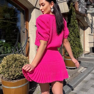 May include: A bright pink knitted two-piece outfit. The top has short, puffed sleeves and a crew neck. The skirt is pleated and falls to mid-thigh. The outfit is worn by a person with dark hair and heart-shaped earrings.