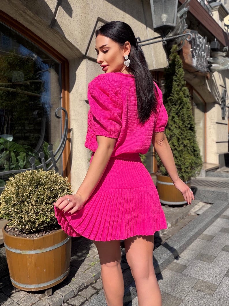May include: A bright pink knitted two-piece outfit. The top has short, puffed sleeves and a crew neck. The skirt is pleated and falls to mid-thigh. The outfit is worn by a person with dark hair and heart-shaped earrings.