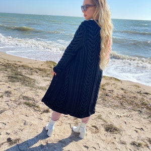 May include: A long, black knit cardigan with a textured, chevron-like pattern. The cardigan is worn over pink pants and white boots. The background features a sandy beach, ocean, and a clear blue sky.