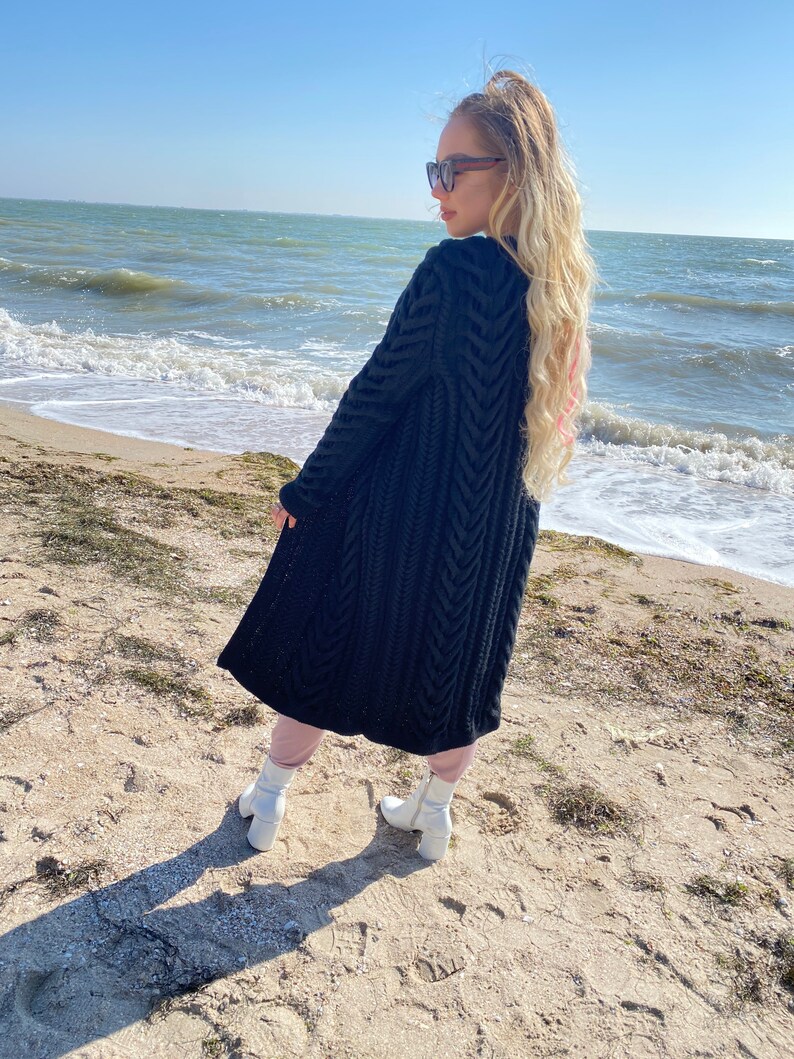 May include: A long, black knit cardigan with a textured, chevron-like pattern. The cardigan is worn over pink pants and white boots. The background features a sandy beach, ocean, and a clear blue sky.