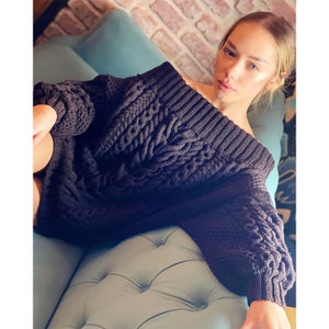 May include: A dark charcoal gray oversized sweater featuring a wide off-the-shoulder neckline and a prominent cable knit pattern. The garment has long, voluminous sleeves with ribbed cuffs and a thick, textured knit construction throughout the body.