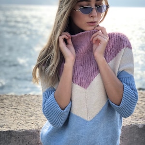 May include: A light blue, white, and pink striped sweater with a high neck and a chevron pattern. The sweater is made of a soft, knitted fabric.