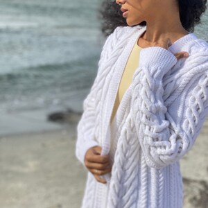 May include: A long, white cable-knit cardigan. The open-front sweater features a textured pattern and long sleeves. The cardigan is worn over a yellow top. The background shows a beach and ocean.
