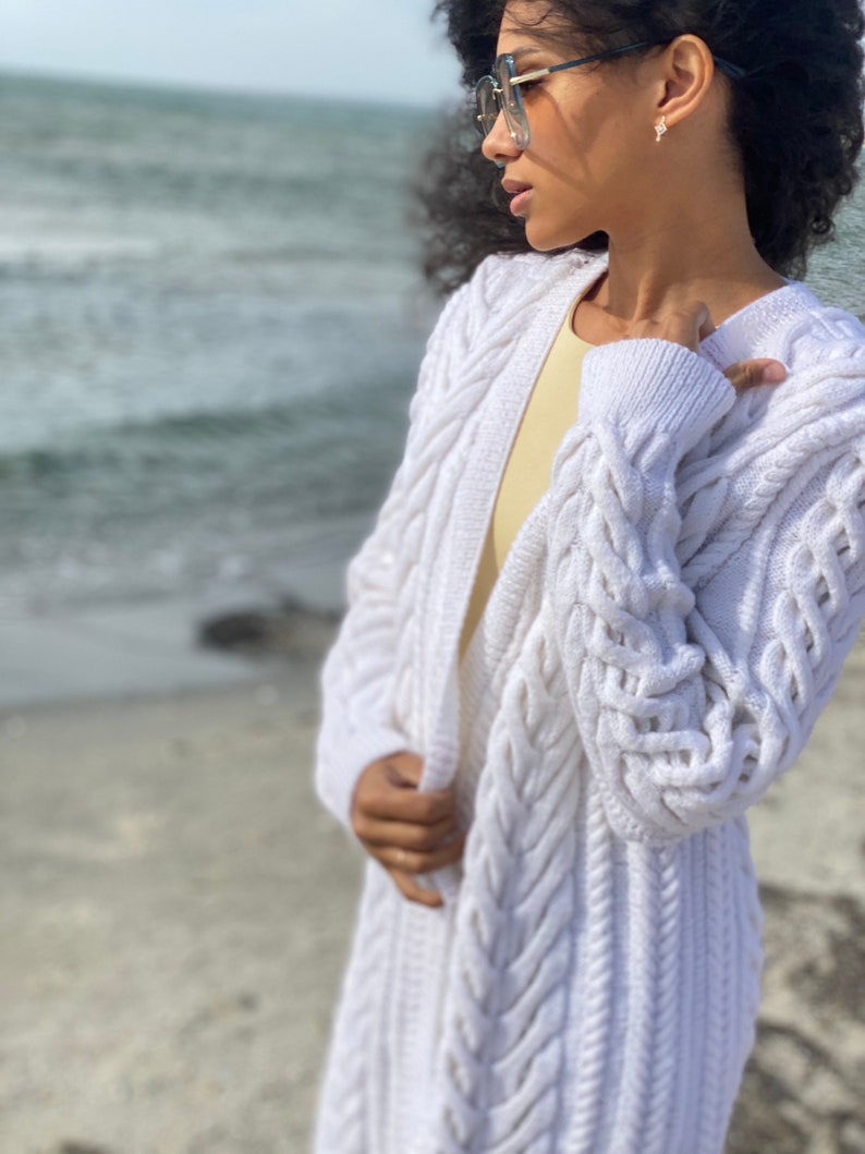 May include: A long, white cable-knit cardigan. The open-front sweater features a textured pattern and long sleeves. The cardigan is worn over a yellow top. The background shows a beach and ocean.