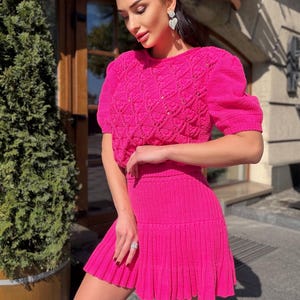 May include: A bright pink knitted two-piece set. The top has short, puffed sleeves and a textured pattern. The matching skirt is pleated. The outfit is a vibrant, eye-catching ensemble.