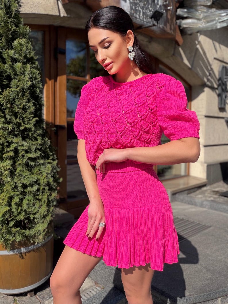 May include: A bright pink knitted two-piece set. The top has short, puffed sleeves and a textured pattern. The matching skirt is pleated. The outfit is a vibrant, eye-catching ensemble.