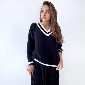 May include: A black sweater with white trim around the neckline and sleeves. The sweater has a v-neck and is worn with a black pleated skirt.