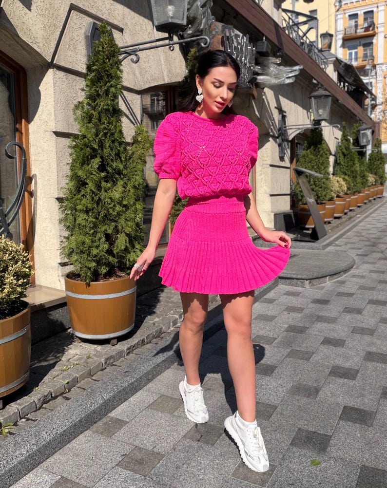 May include: A bright pink knit two-piece outfit, featuring a short-sleeved top with a textured pattern and a pleated mini skirt. The outfit is paired with white sneakers. The ensemble is suitable for warm weather and casual occasions.