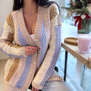 Könnte beinhalten: Ein mehrfarbiger Cardigan mit offenem Schnitt und Zickzackmuster. Der Pullover hat vertikale Streifen in Beige, Hellblau und Hellbraun. Er hat lange Ärmel und ist mit perlenartigen Knöpfen versehen. Der Cardigan ist aus weichem Strickmaterial.