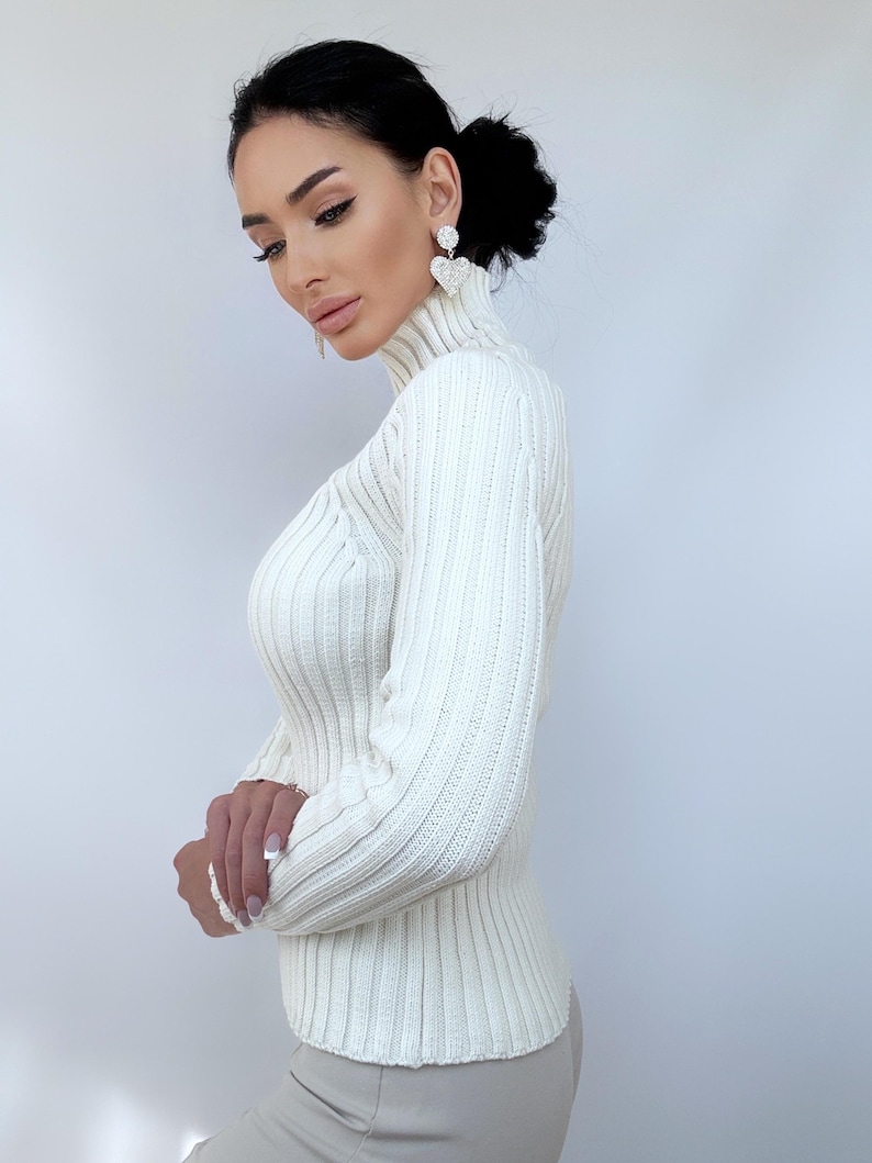 May include: A white ribbed turtleneck sweater with long sleeves. The sweater is fitted and has a high neckline.