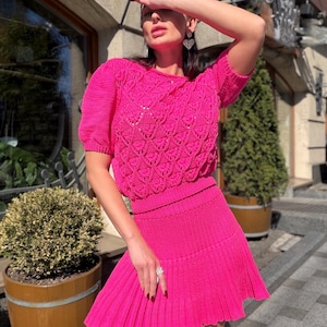 May include: A bright pink, two-piece knit outfit. The top is a short-sleeved sweater with a textured, openwork design. The matching skirt is pleated and mini-length. The outfit is worn outdoors.