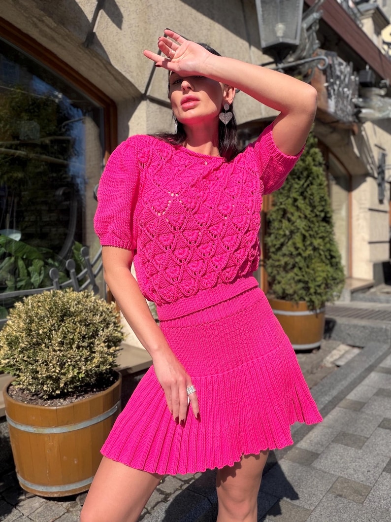 May include: A bright pink, two-piece knit outfit. The top is a short-sleeved sweater with a textured, openwork design. The matching skirt is pleated and mini-length. The outfit is worn outdoors.