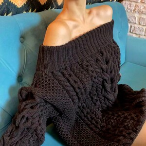 May include: A dark brown off-the-shoulder sweater dress featuring a chunky cable knit pattern on the sleeves and front, combined with a textured honeycomb knit design. The garment has a wide, ribbed neckline that sits across the shoulders.
