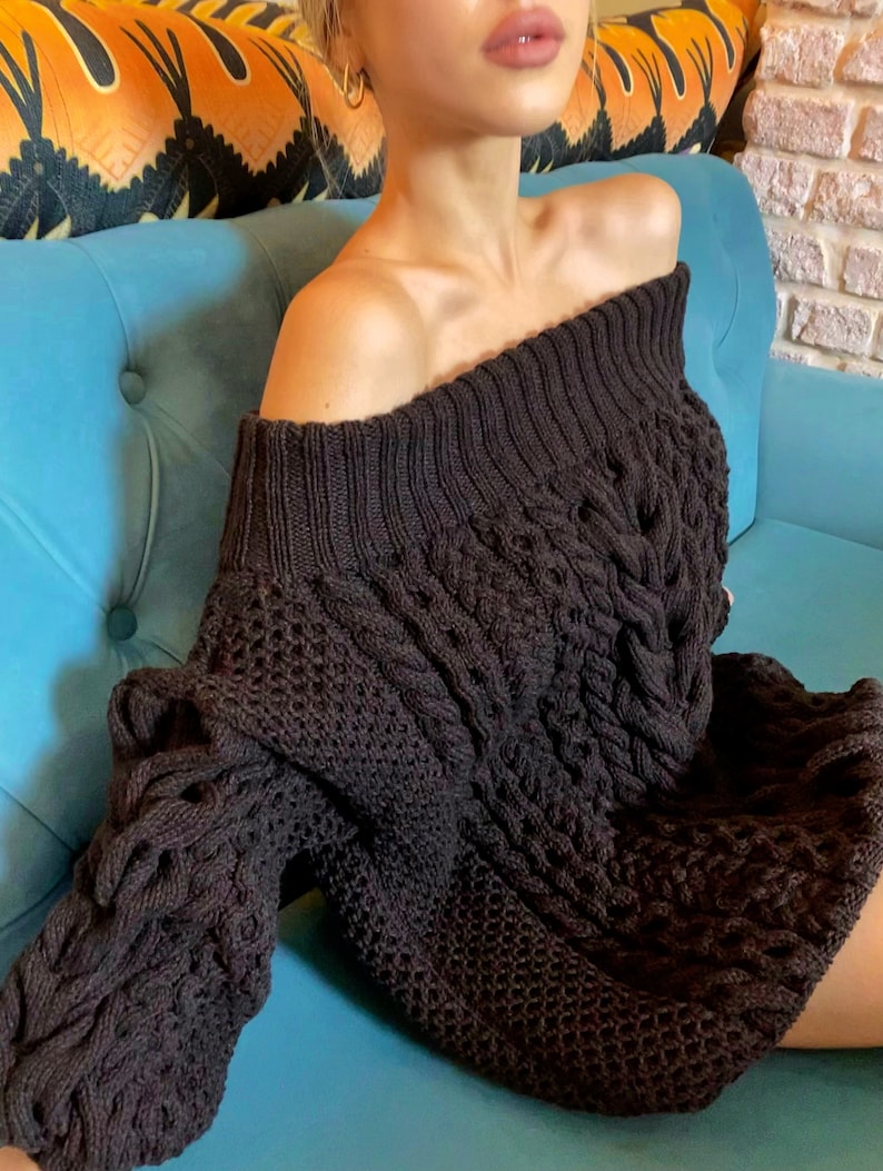 May include: A dark brown off-the-shoulder sweater dress featuring a chunky cable knit pattern on the sleeves and front, combined with a textured honeycomb knit design. The garment has a wide, ribbed neckline that sits across the shoulders.