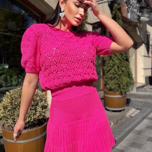 May include: A bright pink knit two-piece set. The top has short sleeves and a textured, open-weave design on the bodice. The matching skirt is pleated and falls just above the knees. The outfit is worn outdoors.