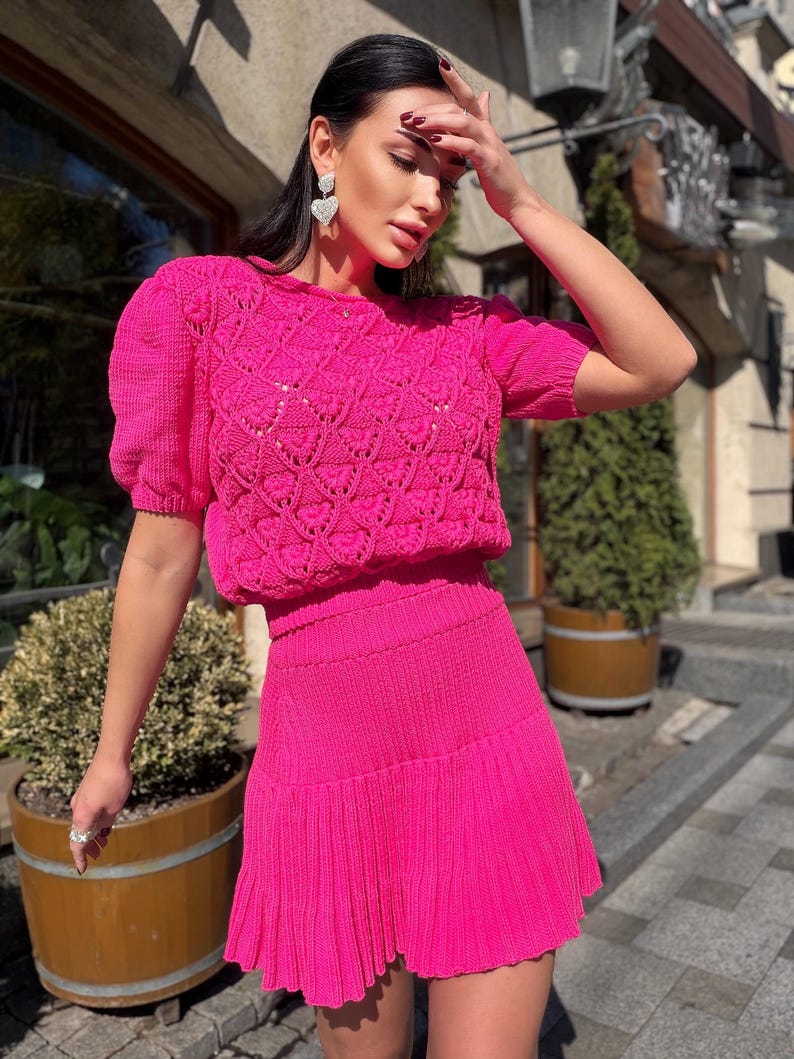 May include: A bright pink knit two-piece set. The top has short sleeves and a textured, open-weave design on the bodice. The matching skirt is pleated and falls just above the knees. The outfit is worn outdoors.