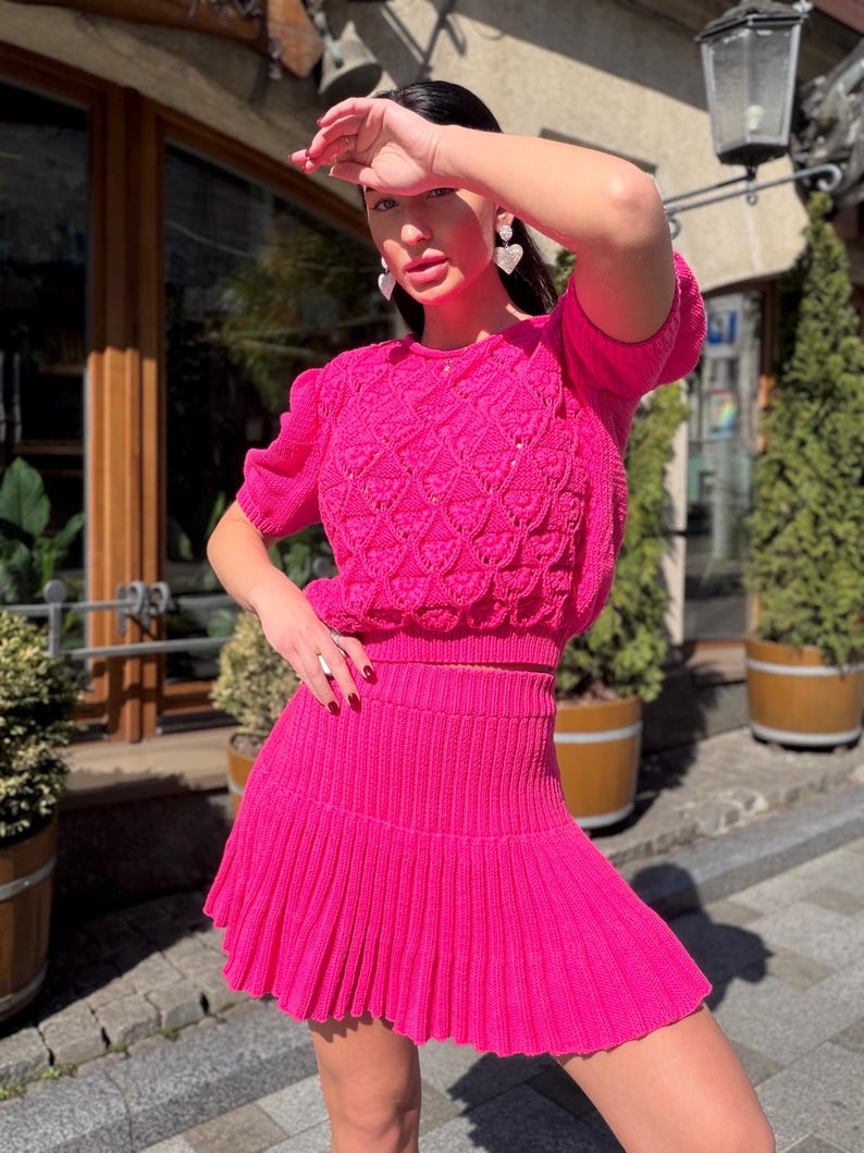 May include: A bright pink knit two-piece set. The top is a short-sleeved sweater with a textured pattern. The matching skirt is pleated. The outfit is worn outdoors in front of a building with trees and a street.