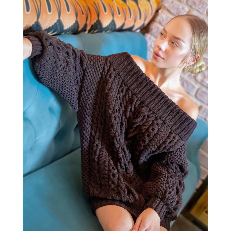 May include: A dark brown, chunky knit sweater featuring an off-the-shoulder neckline, long sleeves with ribbed cuffs, and a prominent cable knit pattern throughout the body. The garment has a wide, ribbed collar and a thick, textured weave.