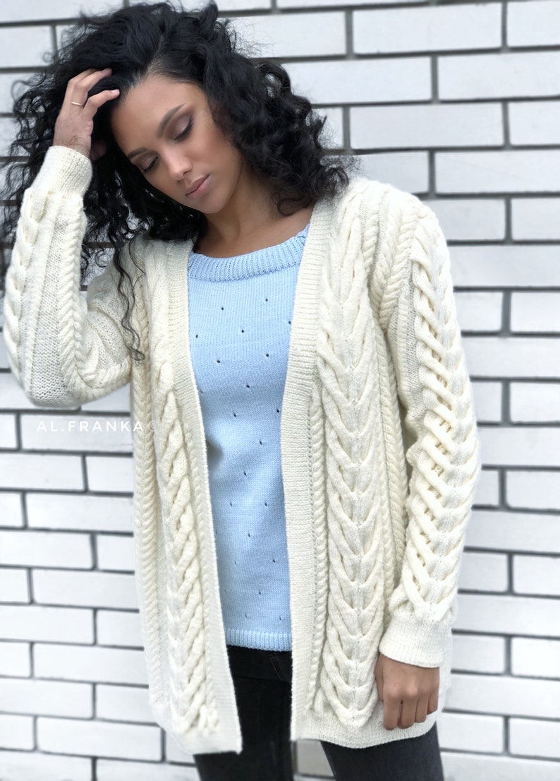 May include: Cream-colored cable knit cardigan with a longline silhouette. The open-front sweater features intricate braided detailing along the sleeves and front panels. A light blue sweater is visible underneath. The background is a white brick wall.