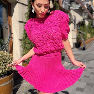 May include: A bright pink knitted two-piece outfit. The top features a textured, open-weave design with short sleeves. The matching skirt is pleated and mini-length. The outfit is a vibrant, eye-catching ensemble.