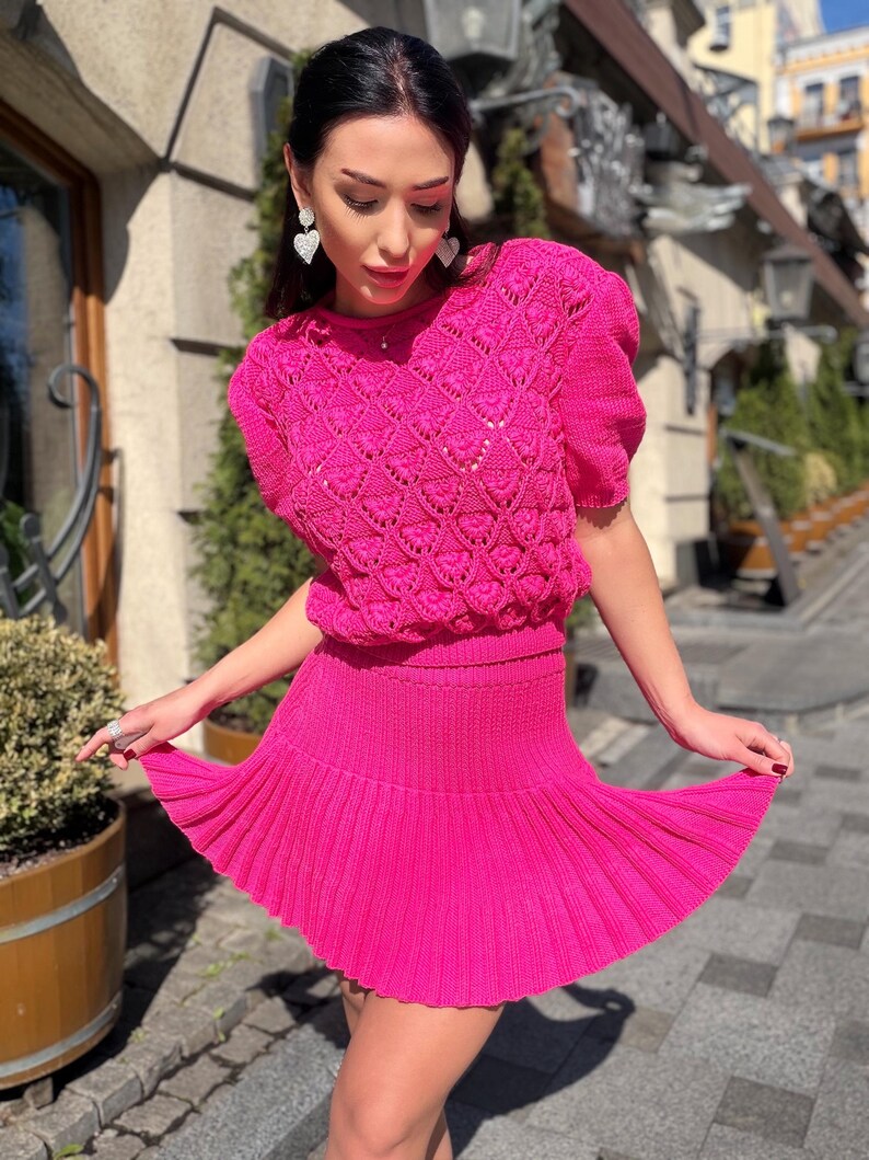 May include: A bright pink knitted two-piece outfit. The top features a textured, open-weave design with short sleeves. The matching skirt is pleated and mini-length. The outfit is a vibrant, eye-catching ensemble.