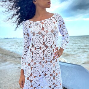K&ouml;nnte beinhalten: Wei&szlig;es H&auml;kel-Strandkleid mit kreisf&ouml;rmigem Muster. Das Kleid hat lange &Auml;rmel und eine lockere Passform.