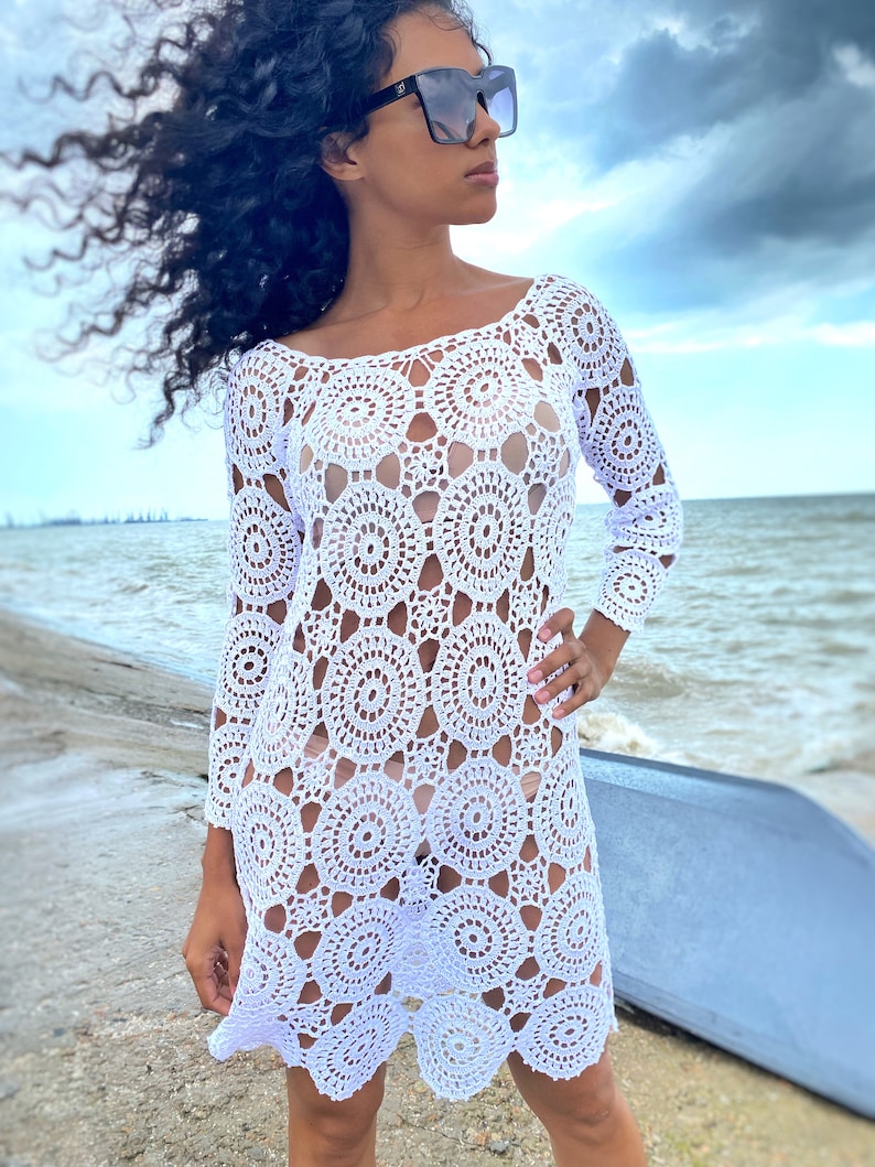 K&ouml;nnte beinhalten: Wei&szlig;es H&auml;kel-Strandkleid mit kreisf&ouml;rmigem Muster. Das Kleid hat lange &Auml;rmel und eine lockere Passform.
