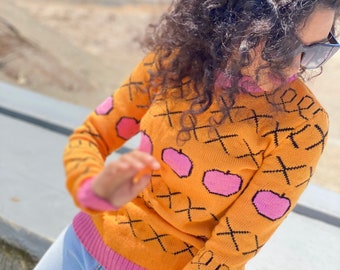 May include: A yellow and pink sweater with a black and white criss-cross pattern and pink apples. The sweater has a pink trim around the neck and sleeves.