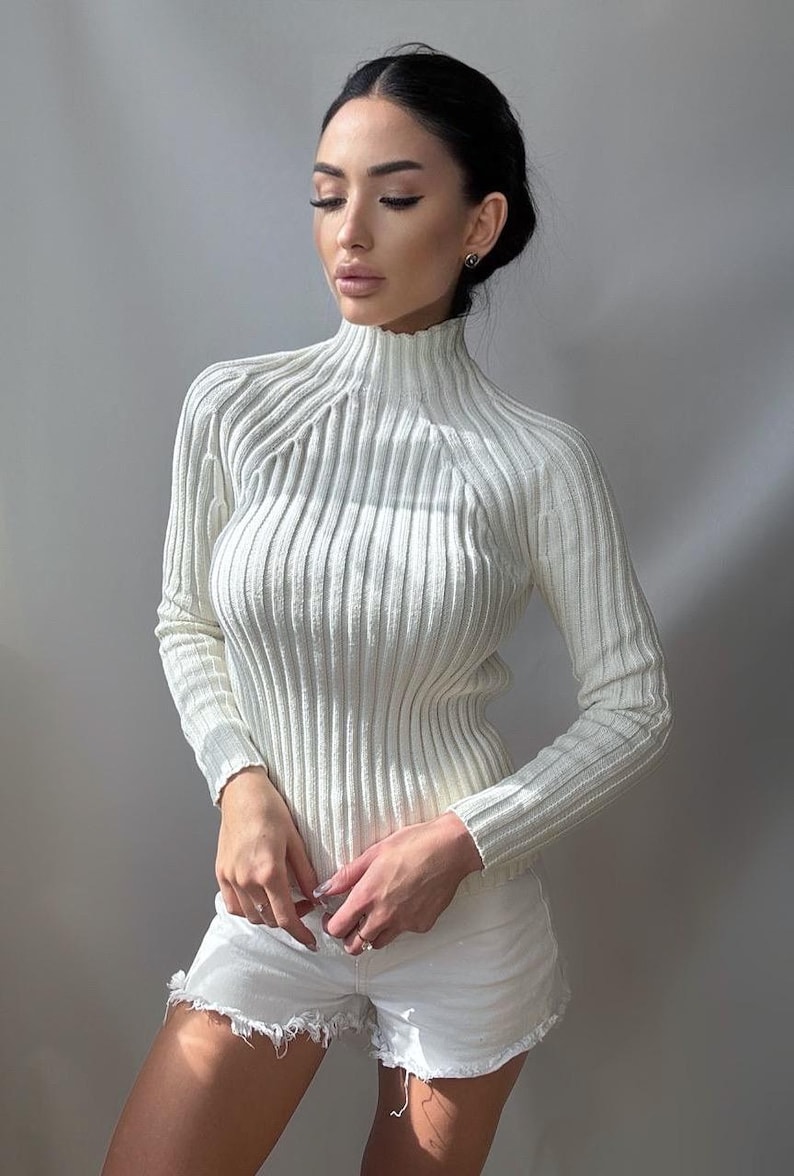 May include: A white ribbed turtleneck sweater with long sleeves. The sweater is fitted and has a mock neck.