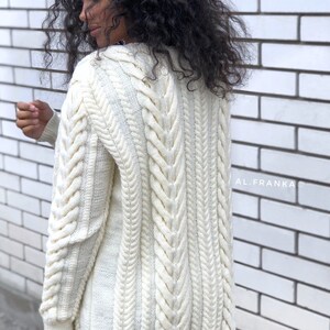 May include: Cream-colored cable knit cardigan with a longline silhouette. The sweater features intricate cable patterns down the sleeves and back, with a ribbed hem and cuffs. The text "AL.FRANKA" is visible on the right side of the image.