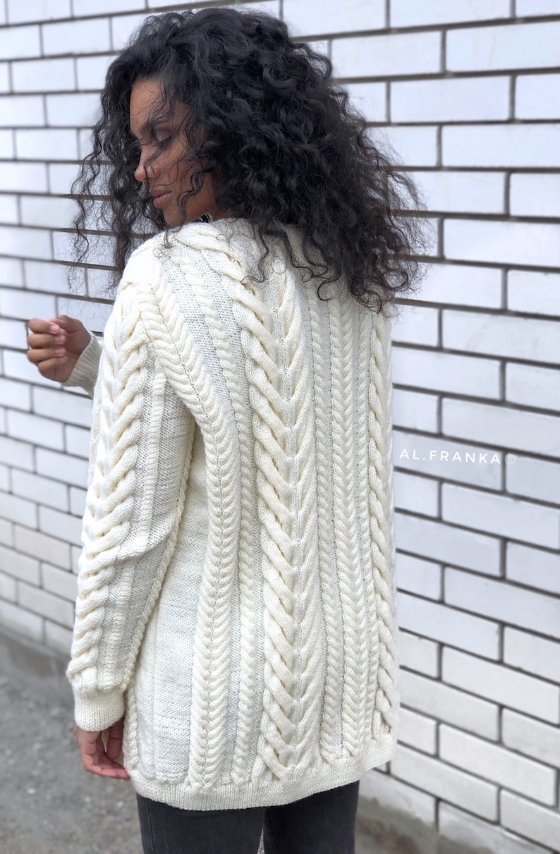 May include: Cream-colored cable knit cardigan with a longline silhouette. The sweater features intricate cable patterns down the sleeves and back, with a ribbed hem and cuffs. The text "AL.FRANKA" is visible on the right side of the image.