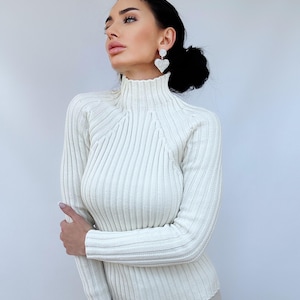 May include: A white ribbed turtleneck sweater with long sleeves. The sweater is fitted and has a high neckline.