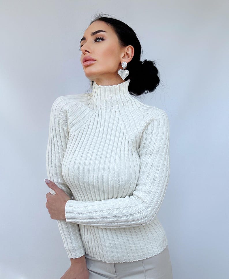 May include: A white ribbed turtleneck sweater with long sleeves. The sweater is fitted and has a high neckline.