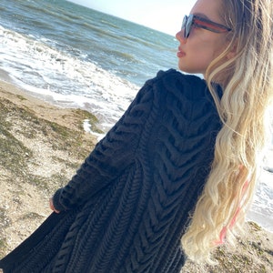 May include: A dark gray, long-sleeved knitted cardigan with a textured, chevron-like pattern. The cardigan is knee-length and appears to be made of a thick, warm yarn. The background shows a beach and ocean.