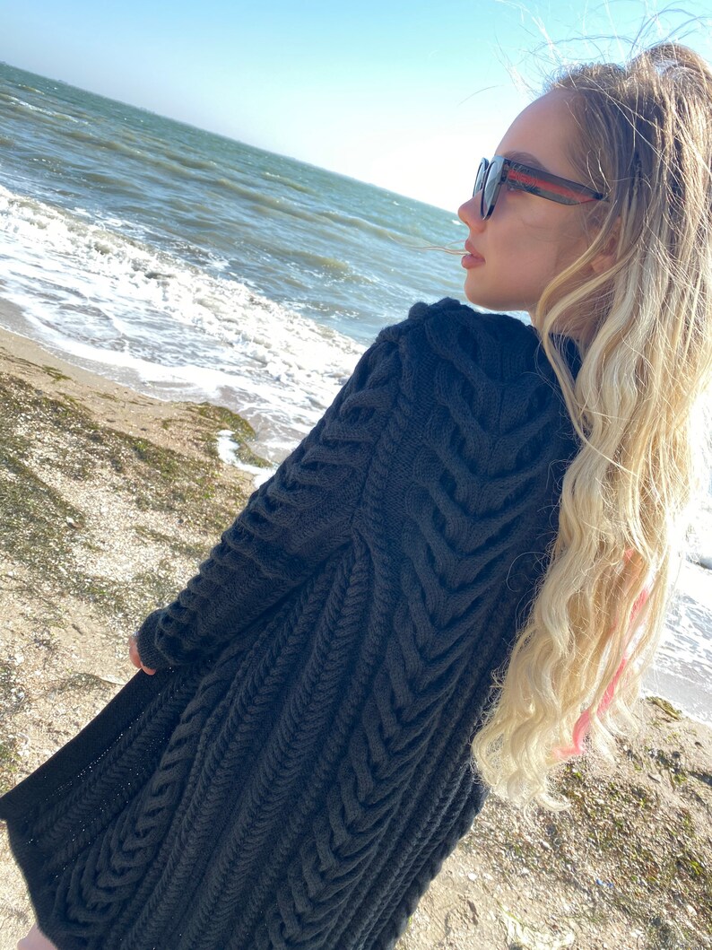 May include: A dark gray, long-sleeved knitted cardigan with a textured, chevron-like pattern. The cardigan is knee-length and appears to be made of a thick, warm yarn. The background shows a beach and ocean.